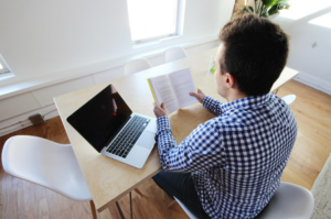 student reading a book while sitting at a desk with a laptop