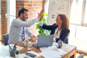 two workers at a table with an open laptop; they are high-fiving each other, a gesture with an upraised arm and palms slapping