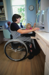 woman in wheelchair working at computer with two children nearby