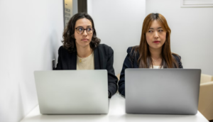 two women at a table with open laptops; women are eyeing each other with expressions of skepticism