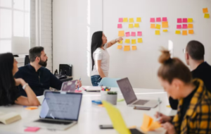 workplace meeting with people and laptops around a table, and a speaker pointing to sticky notes on a wall