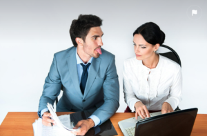two professonals at a desk; one is sticking his tongue out at the other